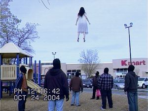 VHS Kmart Levitation Incident — Create a low-resolution VHS camcorder still from 2004 showing a strange paranormal event in a suburban Kmart parking lot beside 