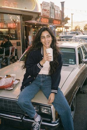 Retro Diner Milkshake Portrait — Create a candid 35mm film-style lifestyle photo of a young woman at a retro American diner during golden hour. She is sitting c