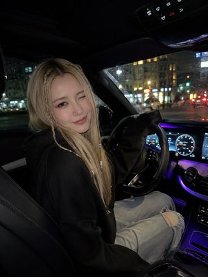 Winking Night Car Selfie — A photorealistic night time car selfie of a young woman sitting in the driver’s seat, turning slightly toward the camera with a playf